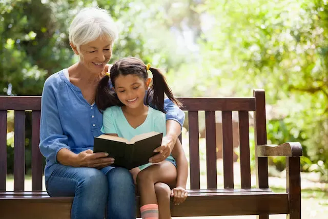 A grandmother reading a book to her granddaughter outside.